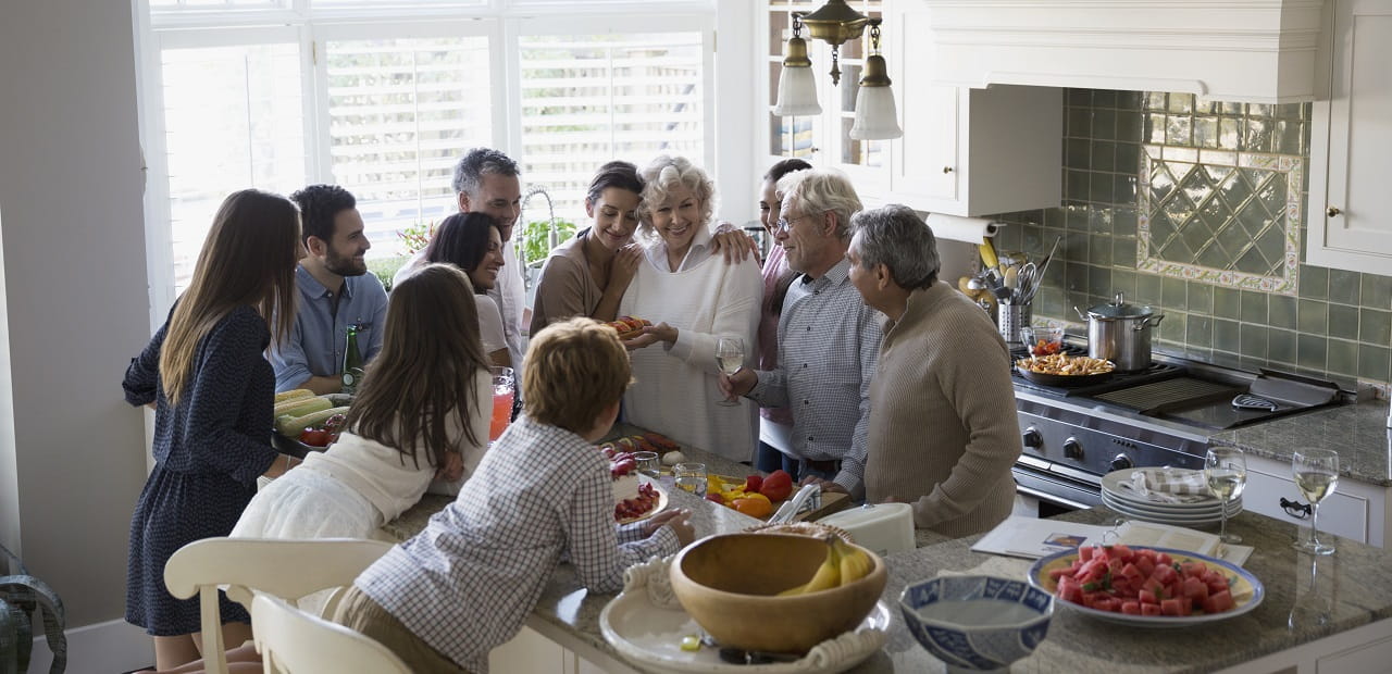 A group of 11 people gathers around a kitchen counter topped with serving plates full of food. 