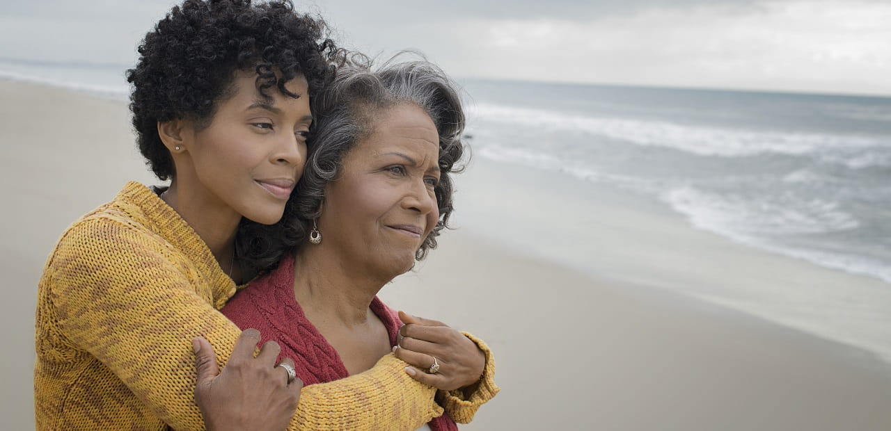 Woman hugs her mother from behind as they look out over the beach. 